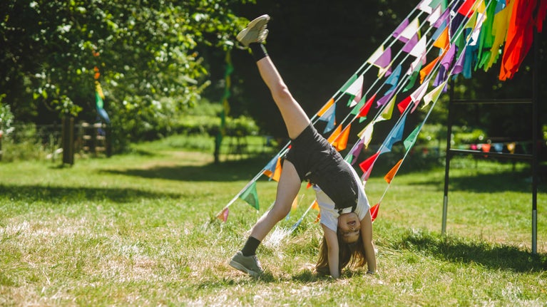 In a grassy field, a child cartwheels in the sunshine, under multi coloured bunting